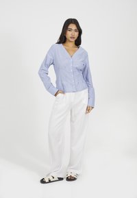 Donna in piedi con una mano in tasca, che indossa una camicetta blu chiaro con bottoni, pantaloni bianchi a gamba larga e sandali bianchi con suole nere.