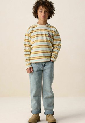 Enfant aux cheveux bouclés portant une chemise à rayures à manches longues, un jean bleu clair avec les revers roulés, et des bottines lacées beige sur un fond neutre.