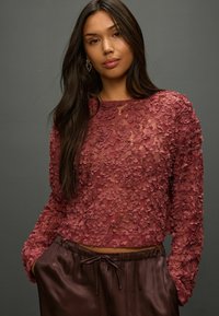 Rose-colored top with textured floral appliqué, long sleeves, and rounded neckline; paired with satin brown pants featuring a drawstring waist.