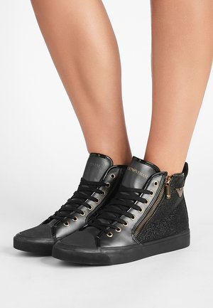 Baskets montantes noires avec œillets dorés, fermetures éclair sur les côtés, panneaux en tissu pailleté et lacets noirs portées sur des jambes nues.