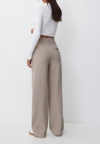 Pantaloni beige a gamba larga con una texture liscia, vita alta e una tasca posteriore singola con dettaglio del bottone nero. Abbinati a una maglietta bianca.