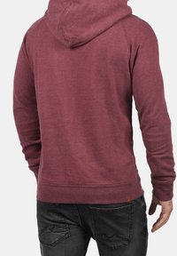 Man i en vinröd huvtröja och svarta jeans, sedd bakifrån mot en neutral bakgrund.