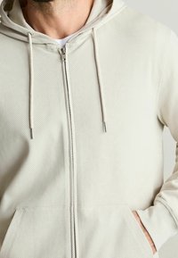 Světlé béžová mikina na zip s texturovanou tkaninou. Má kapuci se stahovací šňůrkou, přední kapsy a doplňky ve stříbrném tónu.