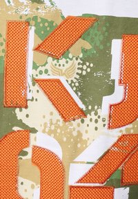 Un tissu en coton présentant un motif camouflage en vert et beige, rehaussé de grandes pièces orange cousues et de formes graphiques blanches.