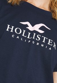 Camiseta de algodón azul marino con un gran logo blanco de "HOLLISTER" y un gráfico de gaviota rosa claro en el frente. Mangas cortas, cuello redondo.