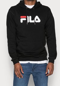 Man i svart FILA-hoodie över vit skjorta och blå jeans, stående med händerna avslappnat vid sidorna mot en enkel bakgrund.