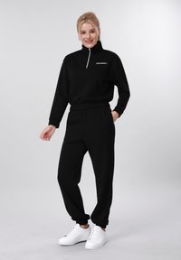 Femme souriante, portant un sweat-shirt noir à fermeture éclair et un pantalon de jogging noir assorti avec des baskets blanches, debout devant un fond clair uni.