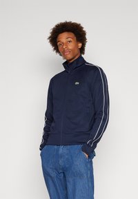 Marineblå glidelåsjakke med høy krage, hvite side-striper og Lacoste-logo. Laget av myk tekstil, har to frontlommer.