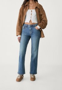 Chaqueta de ante marrón sobre un top corto blanco de canalé con detalles de botones, combinada con unos pantalones vaqueros acampanados de color azul claro y zapatillas marrones.