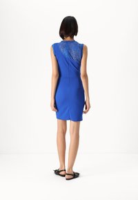 Vestido azul com costas de renda, formato ajustado, até ao joelho, sem mangas, textura suave e uma fenda subtil nas costas. Sandálias planas pretas.