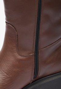 Bottines en cuir marron avec une texture lisse, dotées d'une fermeture éclair noire sur le côté et de coutures renforcées le long des bords. Semelle noire plate.