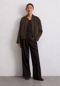 Veste bomber zippée marron avec col montant, associée à un pantalon noir à jambes larges. Textures lisses et silhouette décontractée.