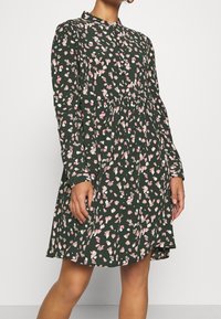 Blumenkleid in Dunkelgrün mit pinken und weißen Mustern, langen Ärmeln, geknöpftem Kragen, gerafftem Taillenbereich und tailliertem Oberteil.