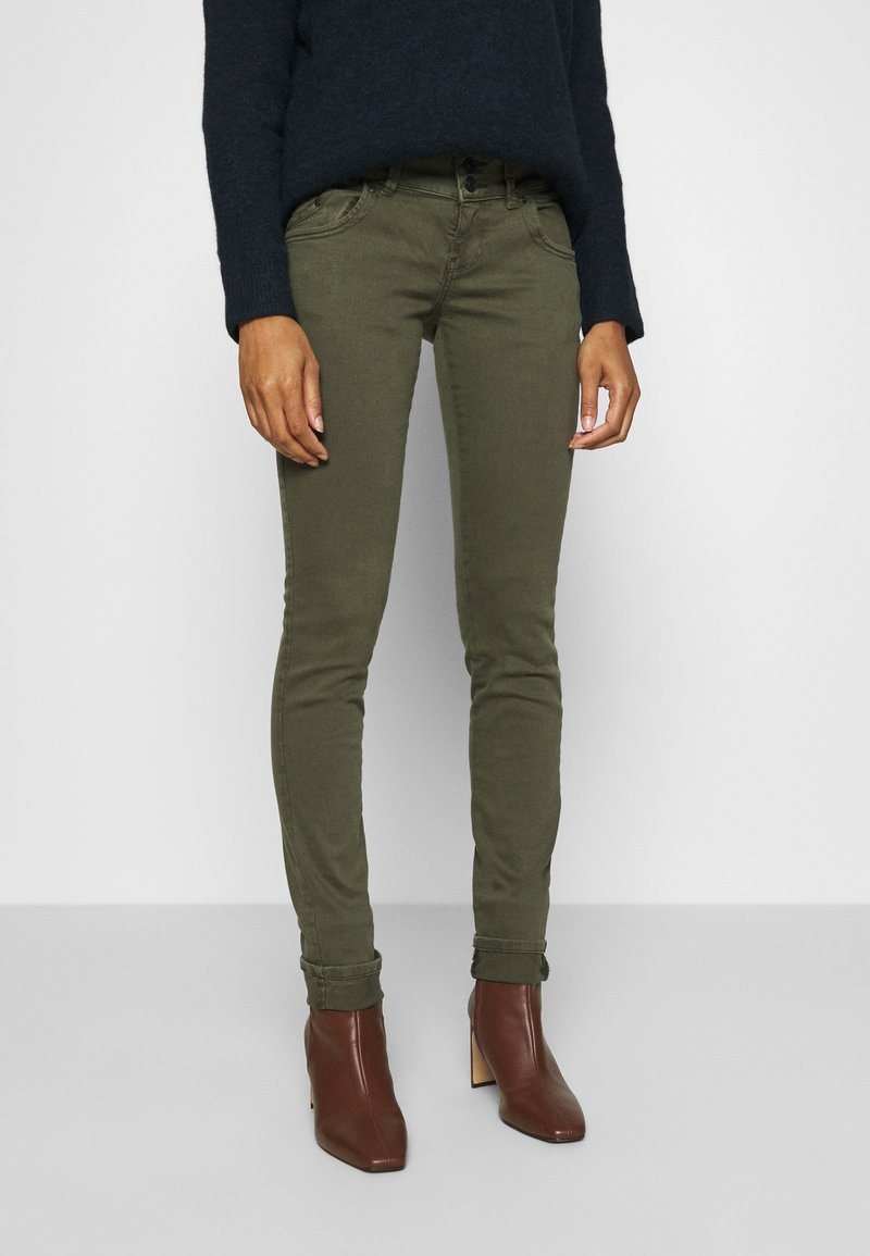 Olivově zelené skinny džíny s měkkou texturou a designem s pěti kapsami, zkombinované s hnědými kotníkovými botami na podpatku. Svetr je tmavě modrý a oversized.