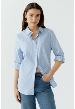 Chemise à rayures bleu clair et blanche avec des manches retroussées, dotée d'embellissements en argent décoratifs. Portée par-dessus un jean en denim.