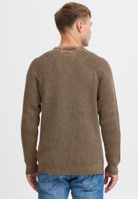 Brązowy, teksturowany sweter z dzianiny z prążkowanymi mankietami i dołem. Posiada metkę na tylnym dekolcie i dopasowany krój. Nosi się go z niebieskimi dżinsami.