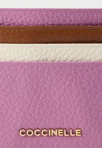 Skórzany portfel z nałożonymi sekcjami w pastelowym różu, brązie i kremowym odcieniu. Złote logo "COCCINELLE" wytłoczone na dole. Teksturowana powierzchnia.