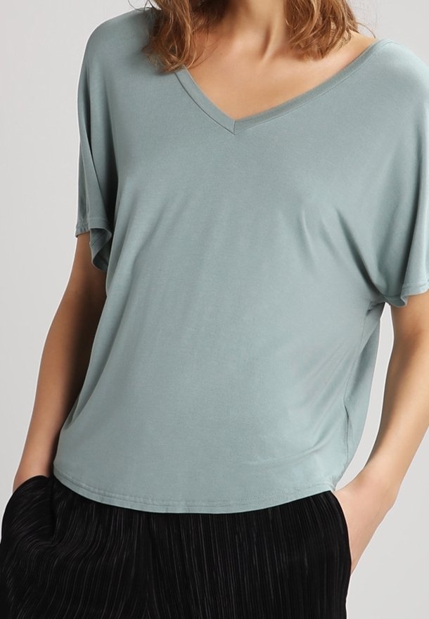 Femme portant un haut ample bleu clair à col en V et manches courtes, les mains dans les poches d'un pantalon noir plissé.