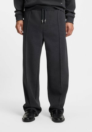 Uomo che indossa pantaloni a gamba larga grigio scuro con cuciture frontali, coulisse nera e scarpe nere, in piedi su uno sfondo neutro.