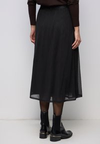 Musta midihame, läpikuultavasta kankaasta, jossa on sileä pinta, istuvalla vyötäröllä ja laskeutuvalla siluetilla. Yhdistetty mustien nilkkureiden kanssa.