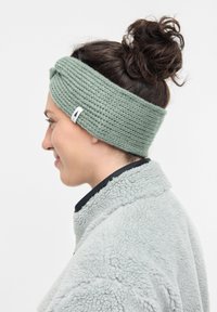 Strickstirnband in sanftem Grün mit gedrehtem Frontdesign. Verfügt über ein kleines weißes Logoetikett. Getragen mit einer hellgrauen Fleecejacke.