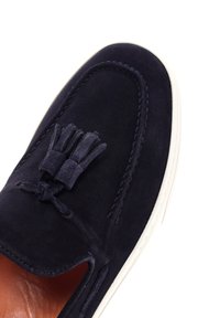 Marineblauer Wildlederschuh mit Mokassin-Design, der eine geknotete Quastenakzente, eine weiße Gummisohle und genähte Details entlang der Kanten aufweist.