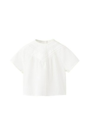 Blouse blanche à manches courtes avec un col montant, un tissu texturé et des détails en dentelle sur la poitrine et les épaules.