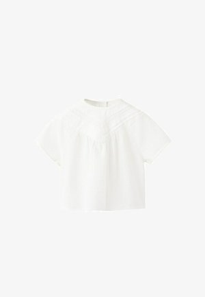 Blouse blanche à manches courtes avec un col montant, un tissu texturé et des détails en dentelle sur la poitrine et les épaules.