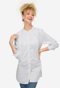 Camicia bianca a maniche lunghe con colletto rotondo e chiusura a bottoni. Presenta un'orlo curvo e un tessuto morbido per il comfort. Indossata con jeans blu.