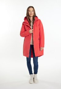 Rød isoleret parka-jakke med hætte, der har lynlås-lommer, trykknapper og en let indsnævret talje, parret med blå jeans og hvide sneakers.