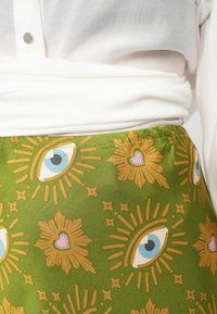 Saia de tecido verde com um padrão que apresenta olhos, corações e elementos florais em tons de dourado e rosa. Textura suave.