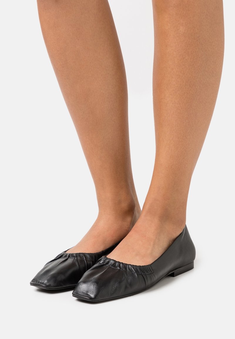 Ballerines en cuir noir à bout carré avec des détails froncés, portées pieds nus sur un fond uni.