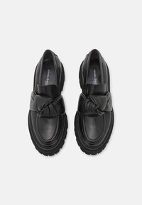 Mocassins en cuir noir avec semelle épaisse et large détail de sangle nouée sur le devant, vus de dessus sur fond blanc.
