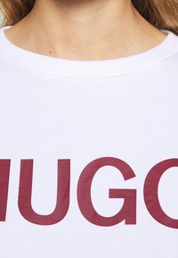 Vitt bomullssweatshirt med ribbad rund hals och stor vinröd "HUGO" grafik tryckt på framsidan. Mjuk känsla, avslappnad design.