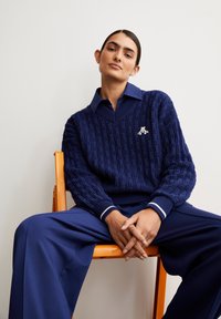 Navyblaue strukturierte Pullover mit V-Ausschnitt, ausgestattet mit weißen Streifen an den Bündchen und einem kleinen Logo auf der Brust, kombiniert mit blauen Hosen.