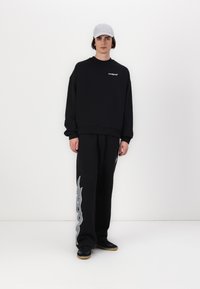 Sort oversized sweatshirt med subtilt logo, parret med sorte bukser med et gråt tentakelmønster, båret med en lys grå kasket og sorte sko.