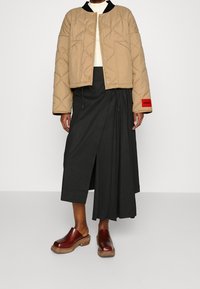 Person iført en beige quiltet jakke, sort asymmetrisk midi-skjørt og brune læder slip-on klipklapper foran en ensfarvet baggrund.