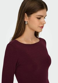 Bordeaux fitted top met een ronde halslijn, lange mouwen en geribbelde textuur. Zilveren druppeloorbellen voegen een subtiele detail toe.