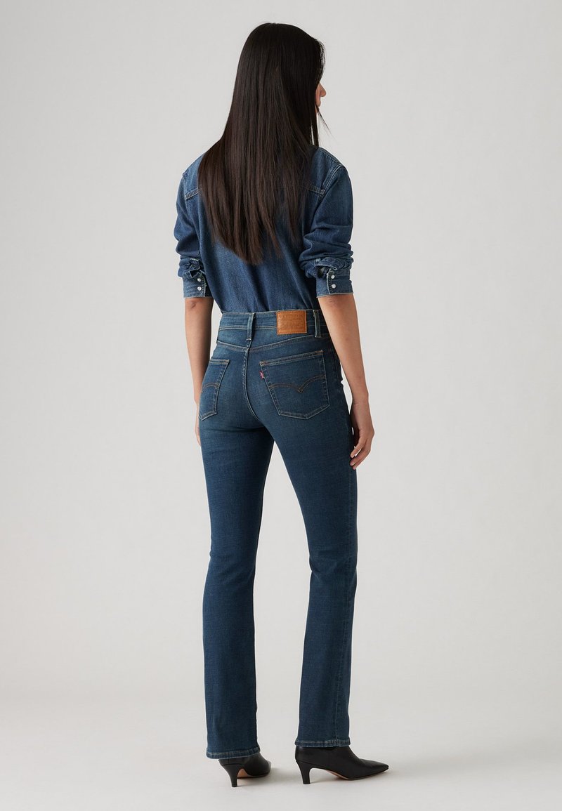 Dunkelblaue, ausgestellte Jeans aus Denim mit hoher Taille, Leder-Patch auf der Rückseite und dezenten Nähten, kombiniert mit einem Denim-Hemd.