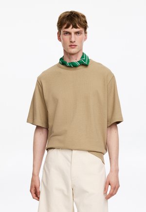 Jeune homme portant une chemise beige oversized à manches courtes, un pantalon blanc et un foulard vert à motifs autour du cou, sur un fond blanc.