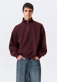 Sweatshirt castanho-avermelhado com gola alta e fecho de meio zíper, apresentando um corte descontraído e punhos elásticos, combinado com jeans azul-claro de perna larga.