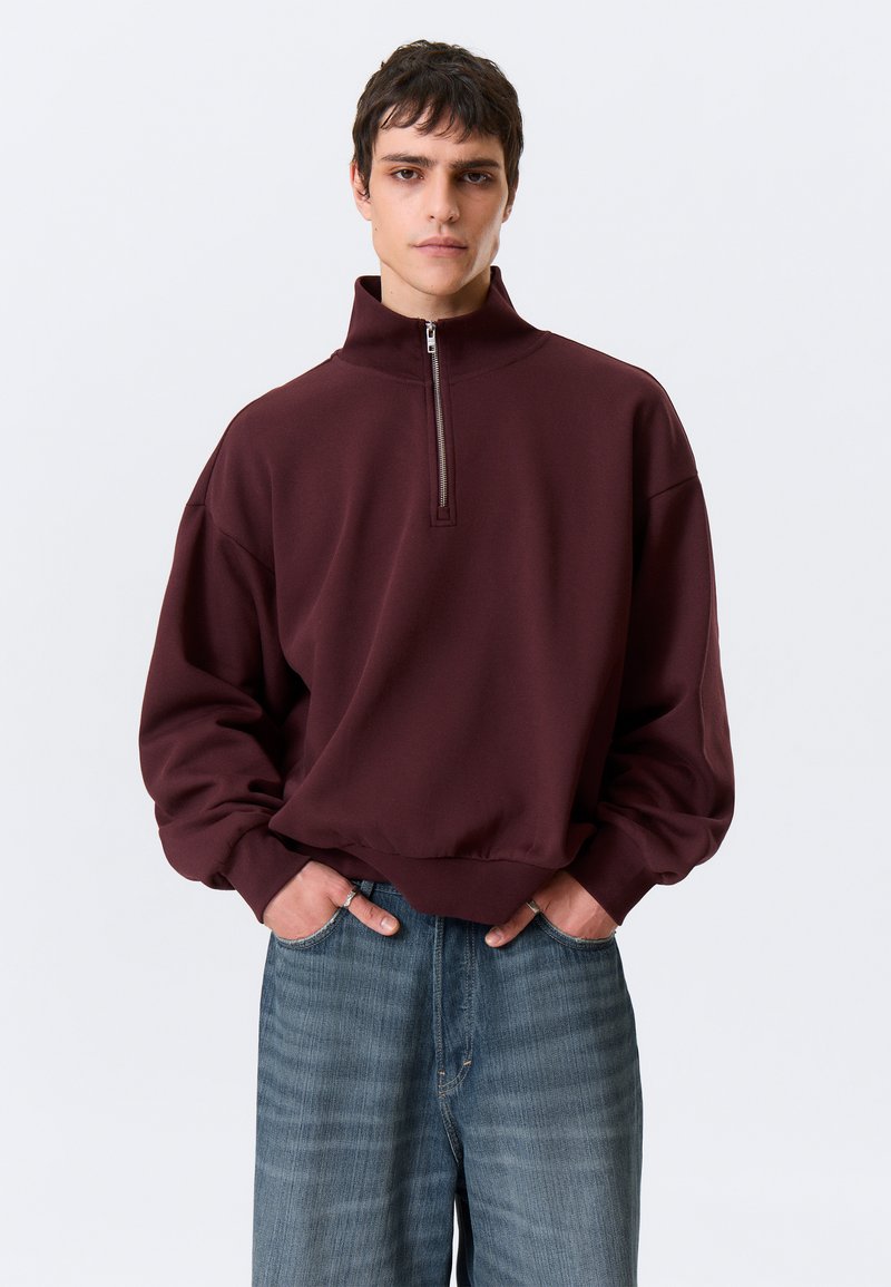 Sweatshirt castanho-avermelhado com gola alta e fecho de meio zíper, apresentando um corte descontraído e punhos elásticos, combinado com jeans azul-claro de perna larga.