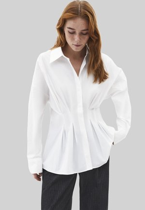 Młoda osoba o długich rudych włosach ubrana w dopasowaną białą koszulę na guziki i ciemne spodnie w prążki, patrząca w dół, z jedną ręką w kieszeni.