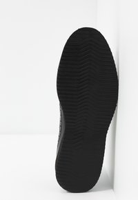 Semelle de chaussure noire avec un motif de bande de roulement épais et texturé, positionnée verticalement contre un fond blanc en coin.
