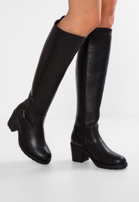 Nogi noszące czarne, skórzane kozaki sięgające do kolan z blokowym obcasem i zamkami z boku, stojące na białej powierzchni na tle jednolitego tła.