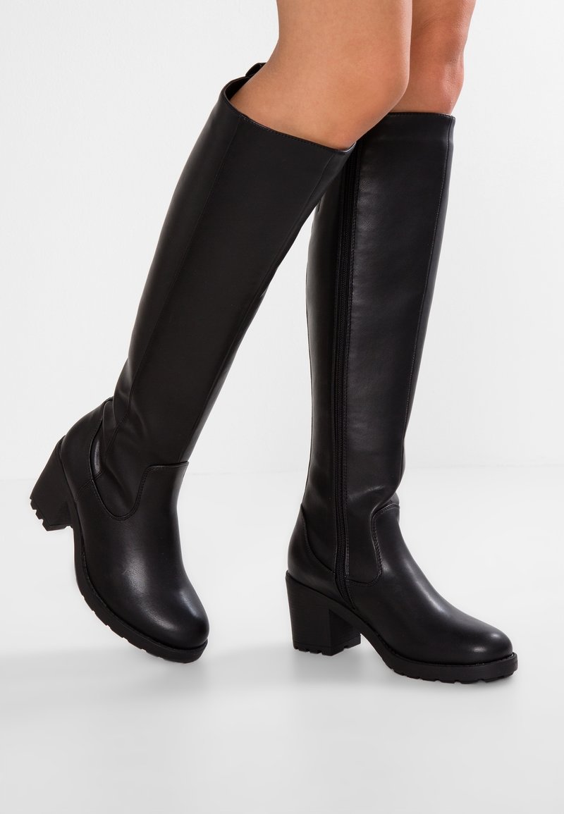 Nogi noszące czarne, skórzane kozaki sięgające do kolan z blokowym obcasem i zamkami z boku, stojące na białej powierzchni na tle jednolitego tła.