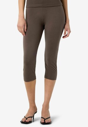 Femme portant des leggings courts marron et des sandales noires à talons, debout sur fond blanc.