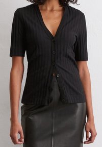 Top negro acanalado de manga corta y botón con un profundo escote en V y cierre frontal de botones, combinado con una falda de cuero negro de cintura alta.