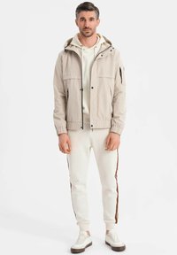 Mann steht da und trägt eine hellbeige Kapuzenjacke über einem cremefarbenen Hoodie, weiße Jogginghosen mit braunen Seitenstreifen und weiße Sneakers mit braunen Sohlen.