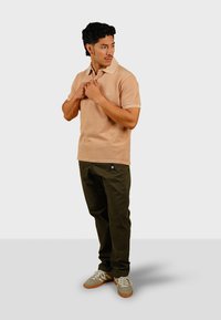 Beige polo shirt met korte mouwen, voorzien van een kraag en geborduurd logo, gecombineerd met olijfgroene broek en lichtgrijze sneakers.
